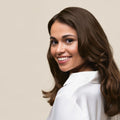 A smiling woman with long, wavy brown hair looks over her shoulder against a plain beige background. She is wearing a white collared shirt.